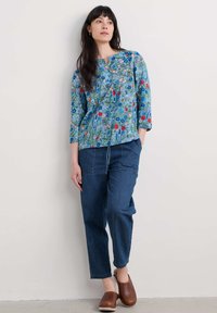 Blouse bleue à motifs floraux avec des manches 3/4 et un col rond, associée à un pantalon en denim foncé ample et des sabots en bois marron.