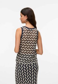 Donna con lunghi capelli scuri che indossa un top senza maniche a zig zag in bianco e nero e una gonna abbinata, di spalle con le braccia piegate.