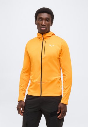 Férfi élénk narancssárga cipzáras dzsekit visel, kis fehér logóval a mellkasán, fekete nadrágban, egy sima világos háttér előtt áll.