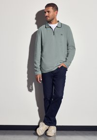 Muž stojící proti bílé zdi, oblečený v světle šedém zipovém pulovru, tmavě modrých džínách a béžových teniskách, s jednou rukou v kapse.