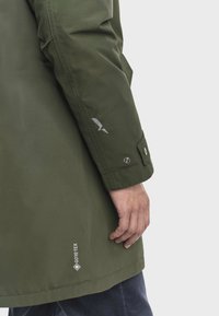 La mano di una persona accanto a una manica di giacca GORE-TEX verde oliva con polsino abbottonato e piccolo logo riflettente sull'avambraccio, su sfondo semplice.