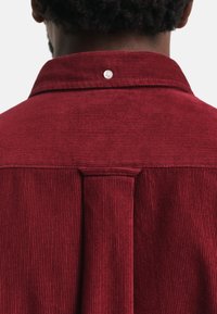 Rode corduroy blouse met een gestructureerd oppervlak, voorzien van een knoop bij de kraag en een subtiele plooi-detaillering langs de achternaad.