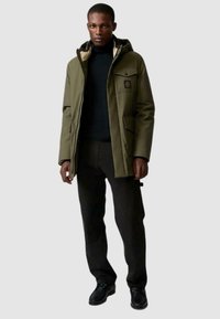 Giacca isolante verde oliva con cappuccio, tasche frontali e rivestimento beige in contrasto, abbinata a pantaloni scuri e scarpe nere.