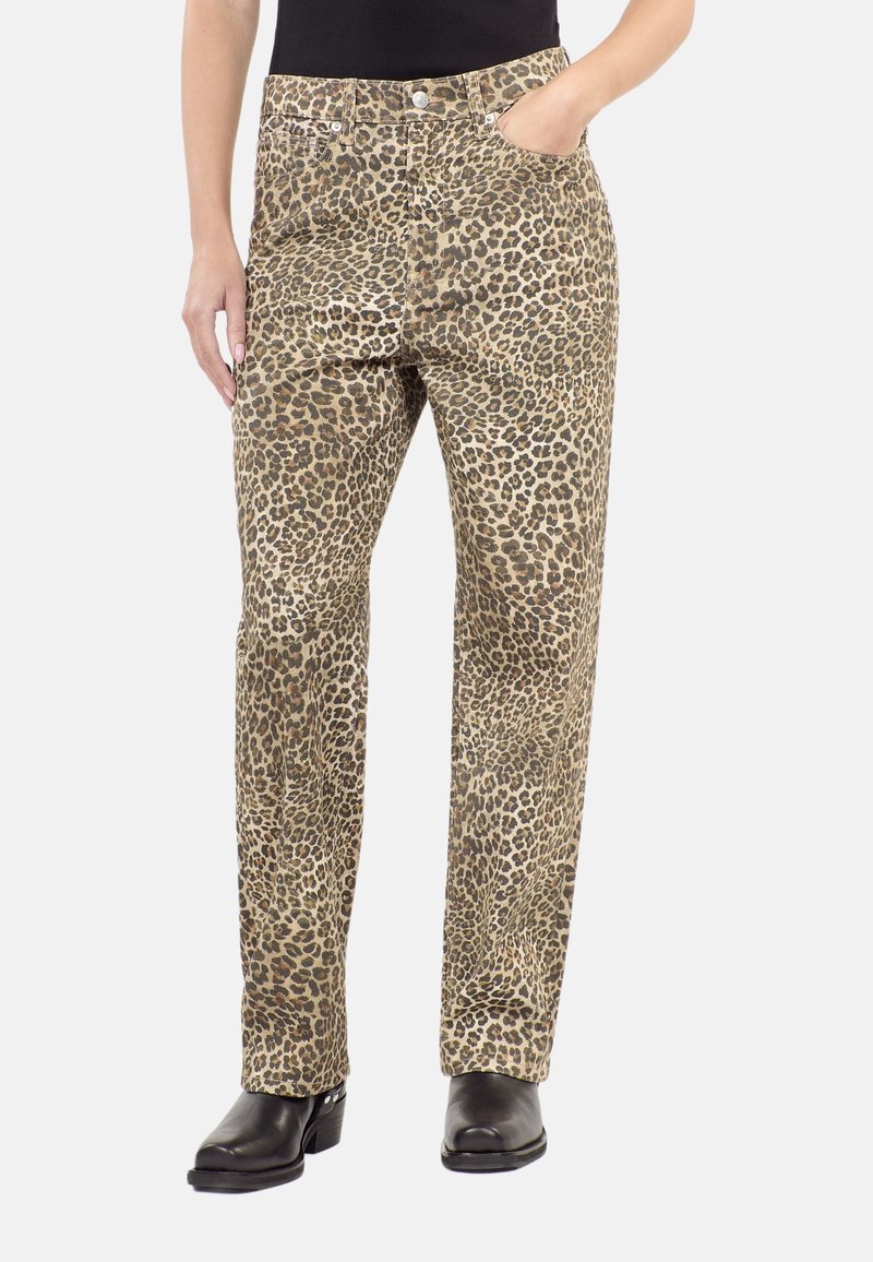 Mujer con pantalones rectos de estampado de leopardo, botas negras, con una mano en el bolsillo y la otra descansando a su lado.