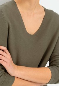 Oliwkowy sweter dziany z głębokim dekoltem w serek, rękawami o długości trzy czwarte i teksturowanym materiałem. Zbliżenie na detal dekoltu i rękawów.