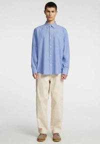 Zila un balta svītraina pogājamā krekla ar garām piedurknēm, kombinēts ar bēšām biksēm un brūnām kurpēm. Audums šķiet gluds un viegls.