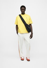 Άνδρας στέκεται με το πρόσωπο προς τα εμπρός, φοράει κίτρινο t-shirt, λευκό παντελόνι φόρμας με ρίγες, κόκκινα αθλητικά παπούτσια και μαύρη τσάντα χιαστί, με φόντο λευκό.