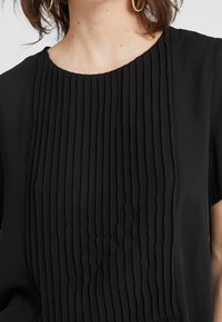 Blouse noire avec des plis verticaux à l'avant, un col rond et des manches courtes, confectionnée dans un textile doux à la texture lisse.