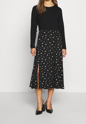 Donna che indossa una maglia nera a maniche lunghe con una gonna midi nera con pois bianchi e beige e uno spacco frontale, abbinata a tacchi neri.
