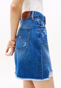 Denim mini skirt in blue with a frayed hem, distressed details, and a brown leather logo patch at the waist. Back pockets visible.