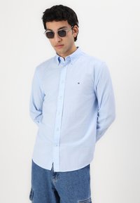 Camisa de botones azul claro con mangas largas y dobladillo curvado. Presenta un pequeño logo cerca del pecho; tejido de algodón suave. Combinada con shorts de mezclilla.