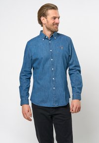 Donkerblauwe denim shirt met button-down kraag, voorste placket en geborduurd logo. Lange mouwen en normale pasvorm. Draagt met zwarte broek.
