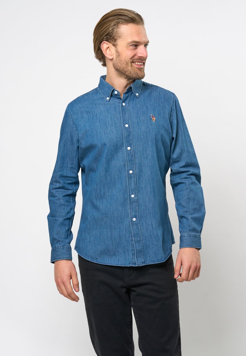 Donkerblauwe denim shirt met button-down kraag, voorste placket en geborduurd logo. Lange mouwen en normale pasvorm. Draagt met zwarte broek.