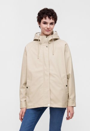 Jeune femme aux cheveux courts et bouclés portant une veste à capuche beige et un jean bleu, debout devant un fond uni.