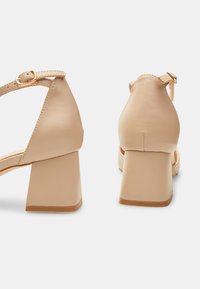 Beige klackskor med slät läderfinish, ankelrem med guldhårdvara och rundad klackdesign.