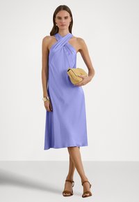 Levanduľové šaty na zaväzovanie okolo krku s prekríženým výstrihom, hladkým textúrou a dĺžkou do polovice lýtok. Modelka drží pletené listové kabelku a má na nohách hnedé sandále na tenkých remienkoch.