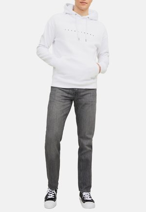 Weißer Kapuzenpullover mit einer Fronttasche und hellgrauem Text. Kombiniert mit dunkelgrauen Jeans und schwarzen Sneakers. Schlichte, lässige Passform.