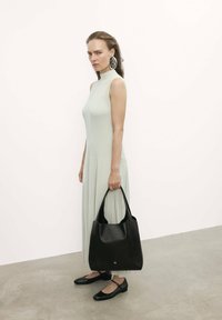 Bolso de cuero negro con acabado texturizado; el modelo lleva un vestido ajustado de canalé verde claro con un cuello alto y zapatos planos negros.