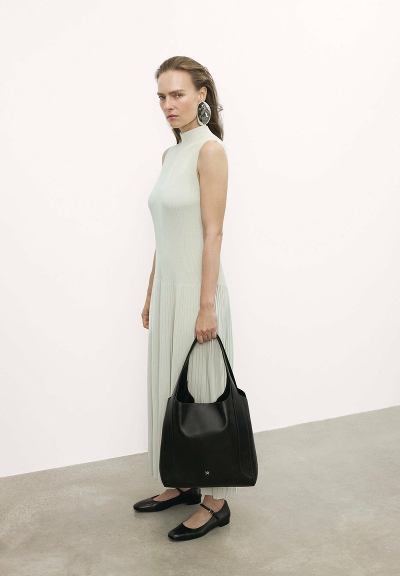 Bolso de cuero negro con acabado texturizado; el modelo lleva un vestido ajustado de canalé verde claro con un cuello alto y zapatos planos negros.