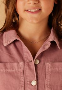 Gouden ketting met meerkleurige kralen, gedragen over een roze spijkerhemd met knoopsluiting en kraag; zichtbare stiksels en een zakdetails.