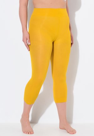 Frau trägt hoch taillierte, leuchtend gelbe kurze Leggings, steht barfuß vor weißem Hintergrund, Hände ruhen seitlich.