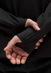 Fekete, hosszú ujjú póló, amelyet illeszkedő mandzsetta, sima textúra és hüvelykujjlyuk jellemez. A kezek hátul helyezkednek el, bemutatva az ujj részletét.