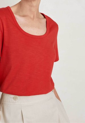 Vrouw draagt een rood T-shirt met korte mouwen en een ronde hals, ingestopt in een high-waisted crème broek met knoopdetail, hand in de zak.