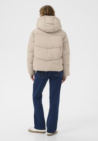 Beige vuorattu takki hupulla, jossa on vaakasaumat ja rento malli. Yhdistetty sinisiin leveälahkeisiin farkkuihin ja valkoisiin lenkkitossuihin.