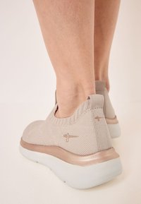 Világos bézs slip-on cipők texturált kötött felsőrésszel, párnázott fehér talppal és fényes rózsaarany díszítéssel az alján.