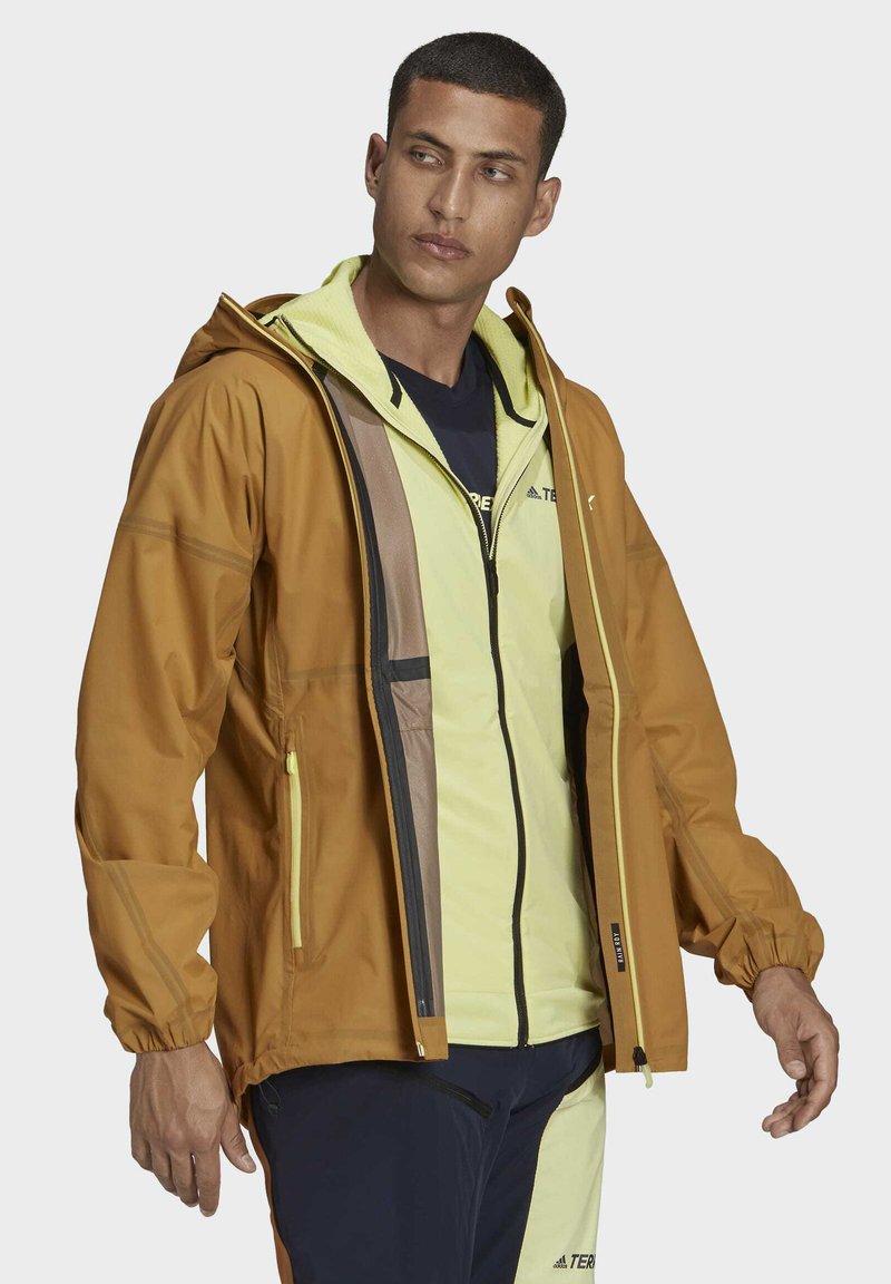 Chaqueta impermeable marrón con capucha, que presenta detalles amarillos, bolsillos con cremallera y un diseño en capas sobre una capa interior amarilla clara.