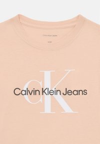 T-shirt pêssego claro com gola redonda, feito de tecido macio, apresentando o texto "Calvin Klein Jeans" em tipografia preta e branca.