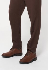 Brązowe skórzane buty z gładkim wykończeniem, okrągłym czubkiem i czarną gumową podeszwą; posiadają sznurowane zapięcie z delikatnymi metalowymi akcentami.