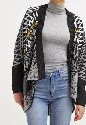Person trägt eine schwarz-weiße und gelb gemusterte Strickjacke über einem grauen Rollkragenpullover und hellblauen Jeans.