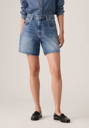 Persoon die midblauwe denim shorts draagt, een denim overhemd in de broek gestopt en zwarte leren loafers, staand tegen een effen achtergrond.
