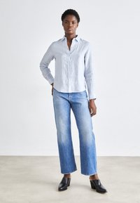 Jasnoniebieska lniana koszula z guzikami, proste, wyblakłe niebieskie dżinsy i czarne mule na obcasie, na minimalistycznym tle.