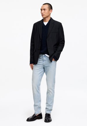 Mężczyzna w czarnej marynarce, granatowym swetrze z dekoltem w serek, białej podkoszulce, jasnoniebieskich dżinsach i czarnych mokasynach, stojący z jedną ręką w kieszeni.