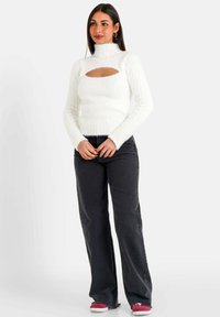 Weißer gerippter Strickpullover mit hohem Kragen und Ausschnitt-Detail. Kombiniert mit schwarzen weit geschnittenen Jeans und Sneakers mit roten Akzenten.