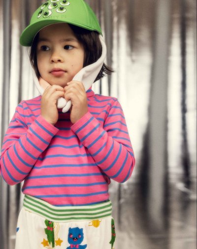 Παιδί που φοράει πράσινο καπελάκι με μάτια, ροζ και μπλε ριγέ πουλόβερ και λευκό παντελόνι με πολύχρωμα καρτούν ζώα, στέκεται μπροστά σε θολό δάσος.