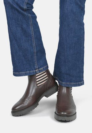 Bottines en cuir marron avec un design brogue, dotées de panneaux élastiques rayés et de semelles en caoutchouc texturé, associées à un jean en denim.