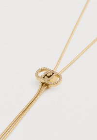 Zlatý náhrdelník s obdĺžnikovým uzáverom zdobeným malými čírymi drahokamami na svetlom pozadí.