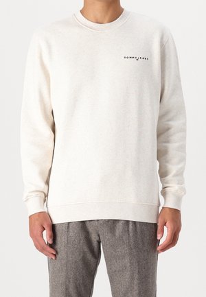 Mann trägt einen off-white langärmligen Pullover mit kleinem "Tommy Jeans"-Logo auf der linken Brustseite und grauen strukturierten Hosen.