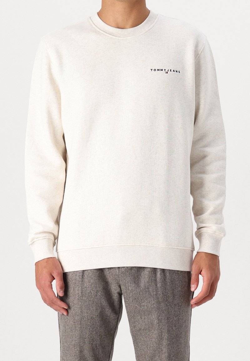 Homme portant un sweat-shirt à manches longues écru avec un petit logo "Tommy Jeans" sur le côté gauche de la poitrine et un pantalon gris texturé.