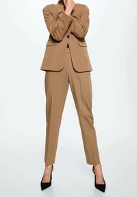 Persona che indossa un completo marrone su misura con giacca abbottonata e pantaloni, in piedi con una mano vicino al viso e tacchi alti neri.