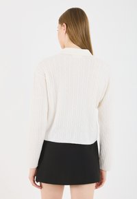 Λευκό πουλόβερ με πλέξη καναπέ σε cropped σχέδιο και μακριά μανίκια, συνδυασμένο με μαύρη μίνι φούστα. Διατηρεί μια ομαλή, υφασμάτινη εμφάνιση.