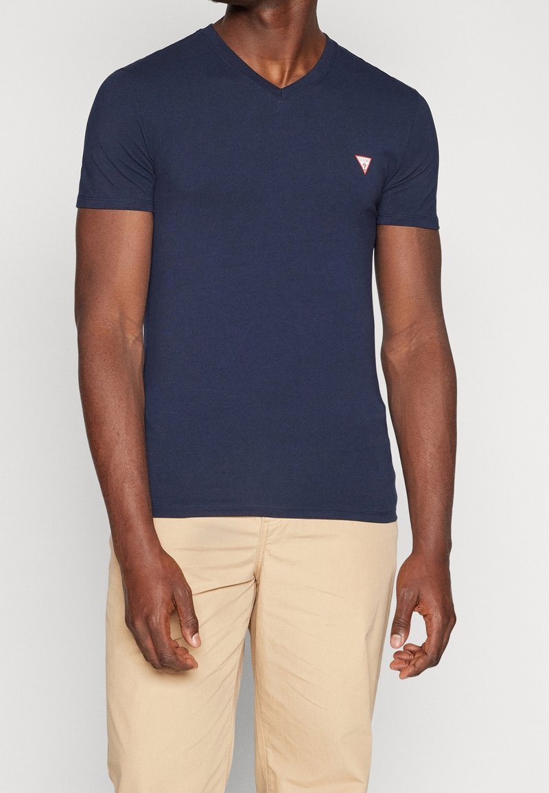 Homme portant un t-shirt bleu marine ajusté à col en V avec un petit logo triangulaire sur la poitrine et un pantalon beige, debout devant un fond uni.