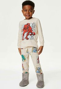 Komplet piżam dla dzieci z kremową bluzką na długim rękawie z czerwonym wzorem graficznym oraz beżowymi spodniami z kolorowymi wzorami superbohaterów. Szare, puszyste kapcie.