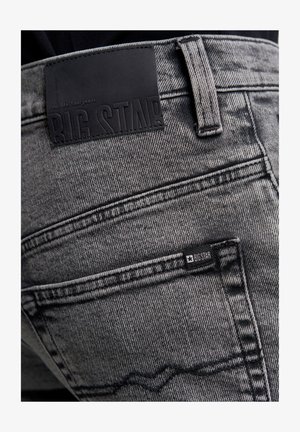 Mustad hallid teksad, millel on tekstureeritud viimistlus, suur nahklabel ja väiksem brändi silt tagakoti lähedal.