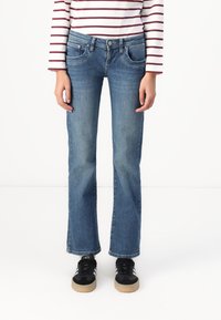 Vaqueros de mezclilla azul con corte recto, que presentan bolsillos delanteros y un cierre de botón. Combinados con una blusa de rayas de manga larga y zapatillas negras.