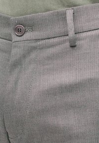 Gros plan sur un pantalon gris à motif chevrons avec un bouton, une passoire de ceinture, une ceinture cousue et le bord partiel d'une poche visible.