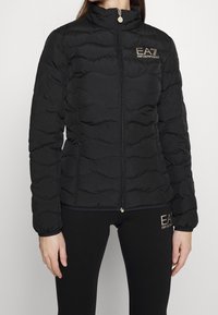 Czarna puchowa kurtka z wysokim kołnierzem, pikowana, z przednim zamkiem błyskawicznym i złotym logo. W komplecie czarne legginsy z podobnym logo.